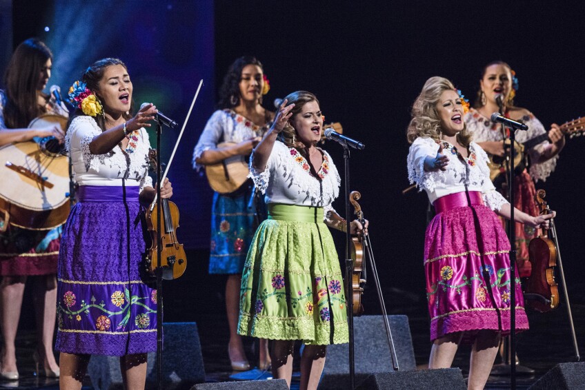 Three women wearing brightly colored skirts with embroidered flowers each stand behind a microphone as they sing, holding their hands up as they sing into the mic. With their other hands, they are each holding a violin and bow. Behind the three women are three other women, wearing similar costuming, but playing guitars.