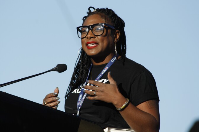 A woman with braided hair, red lipstick and thick, black sunglasses speaks at a podium.