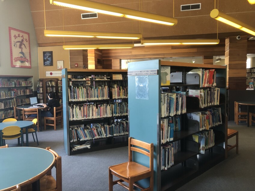 Rows of green-blue colored book shelves display various books inside the Los Feliz Branch Library. Above the bookshelves are yellow hanging pendant lights. 