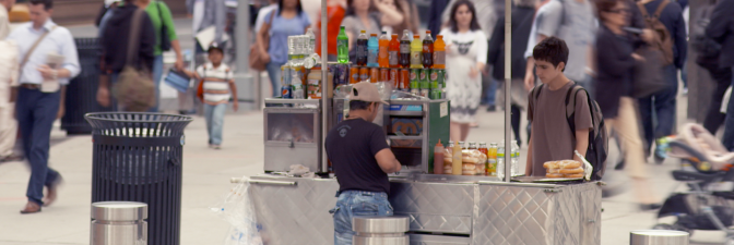 A young customer waits for his order at a hot dog stand on a busy street. | Still from "City Rising: Informal Economy"