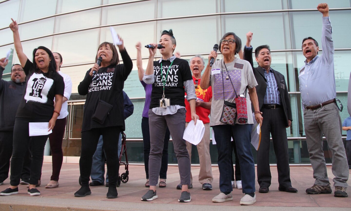 About ten people stand together speaking into microphones. Several raise their fists to the sky.