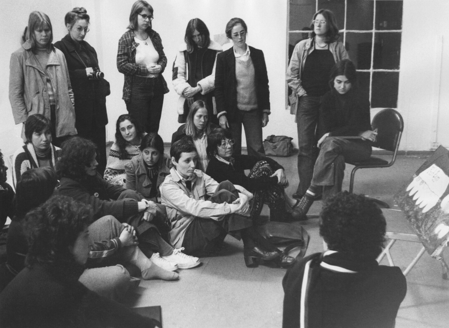 Some women sit on the floor, others stand, all are looking at a painting on a chair.