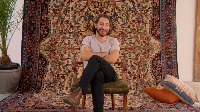 Ari, a man with shoulder-length dark brown hair and a beard, sits on a green velvet upholstered chair with his legs crossed and his arms folded across his chest. He is sitting relaxed as he laughs and looks at something behind the camera. The backdrop is an ornate rug hanging from the ceiling and curtains on the floor. Two accent pillows sit casually to the side and a potted palm tree stands to the side.
