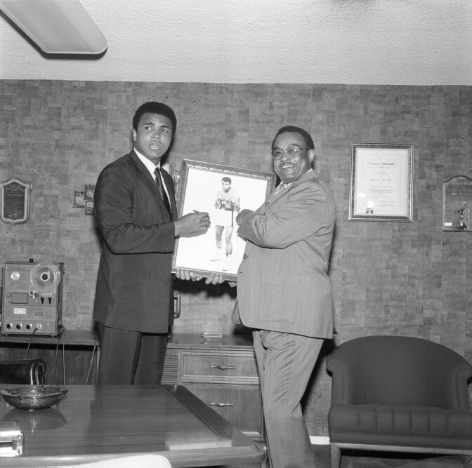 Two African American men stand with a photo frame in between them.