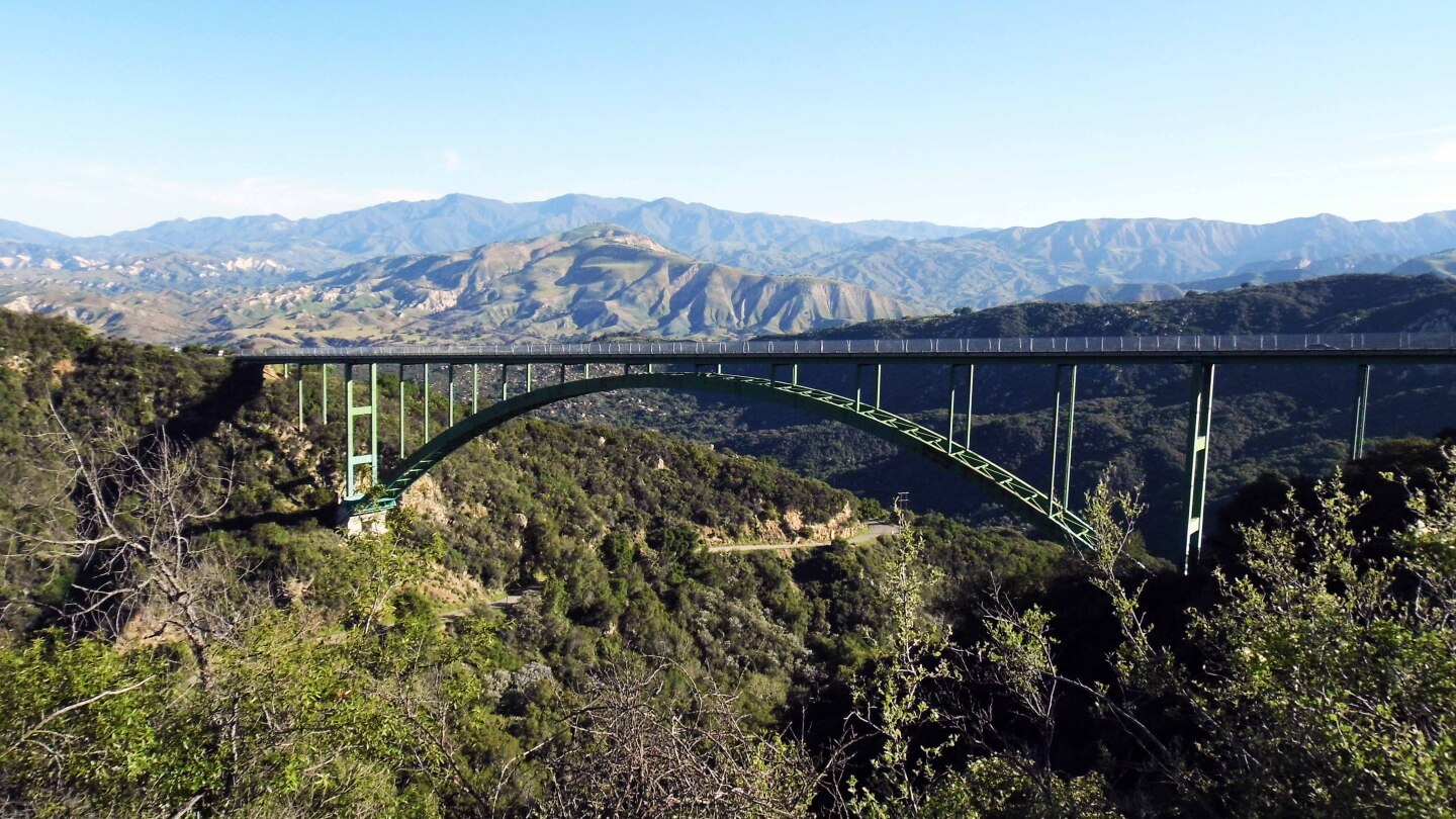 A green arch bridge made of metal beams stretches between two mountains. Behind it are rolling mountains and a clear blue sky.