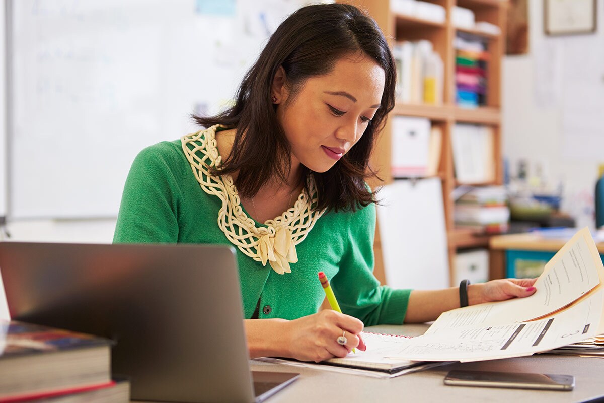 A woman in a green sweater looks down at some papers as she holds a pen. A laptop can be seen next to her. istock