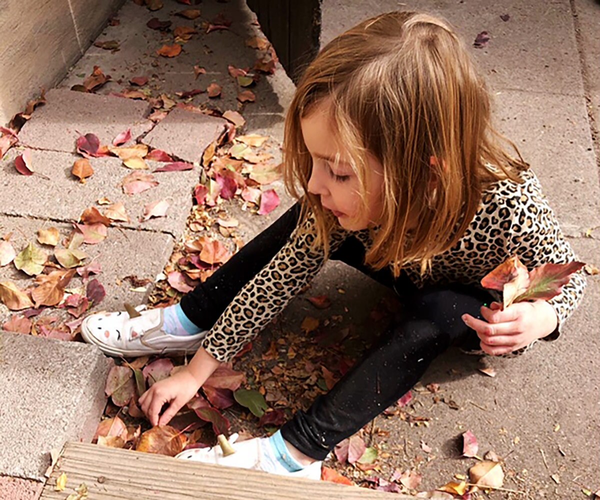 A small blonde girl sits on the floor gathering leaves