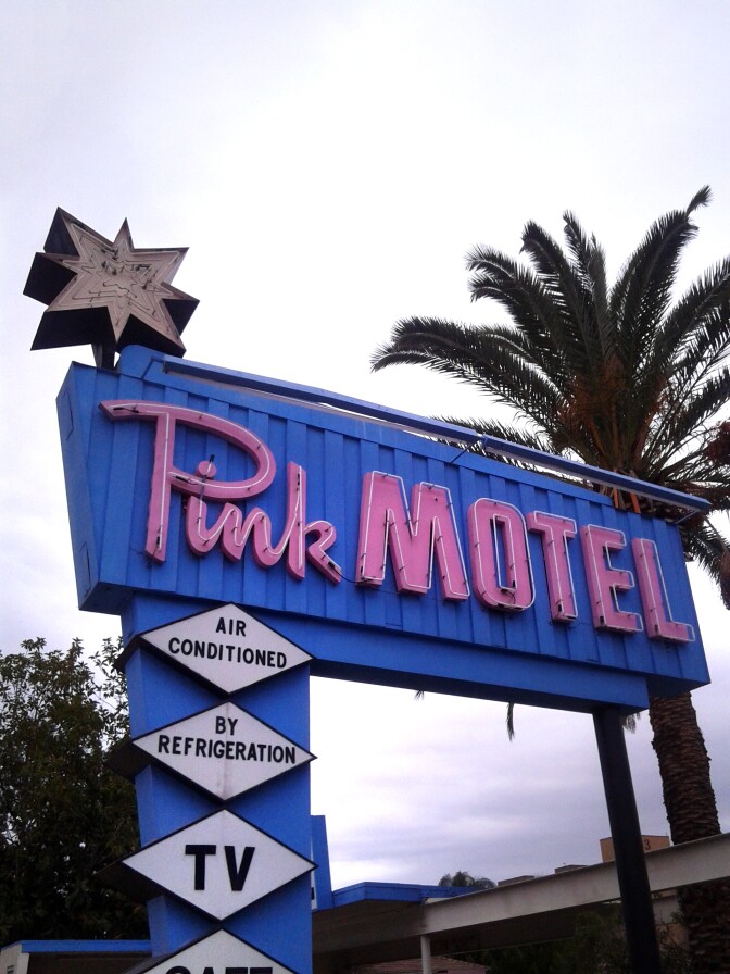 A mid-century neon smotel sign reads, "Pink Motel" in bright pink lettering over a electric blue background. Along the post holding up the sign are diamond-shaped attachments that read, "Air conditioned," "by refrigeration."