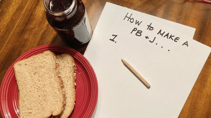 Overhead shot of the making of a peanut butter and jelly sandwich . There is a sheet of paper that reads "how to make a pb+j ... step 1," indicating that the steps should be filled in.