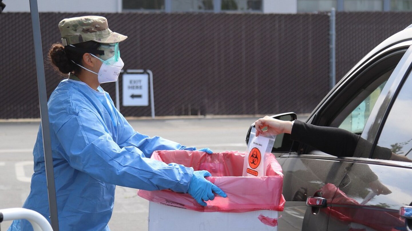 A member of the national guard in blue protective gear holds a box out to a person placing a coronavirus test in a box from a car.