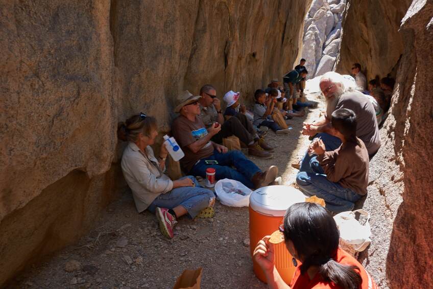 A group of men, women and children sit in a narrow passageway in between two large rock formations. They sit on either side of the pathway as they eat and drink together.