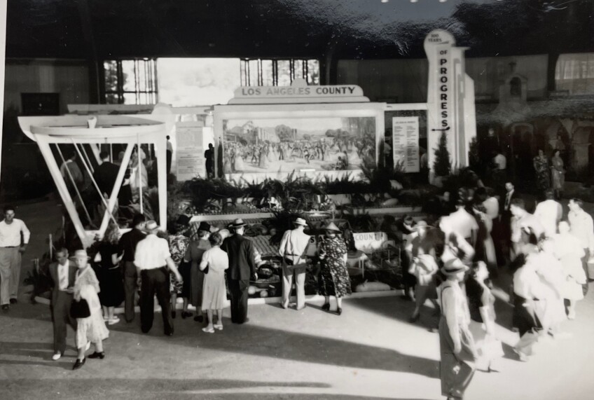 A black and white photo of attendees gathered around an exhibit. The main centerpiece of the exhibit is a large sign with artwork of old Los Angeles County. Above the artwork is a sign that reads, "Los Angeles County." Attendees look on at the exhibit from all sides.