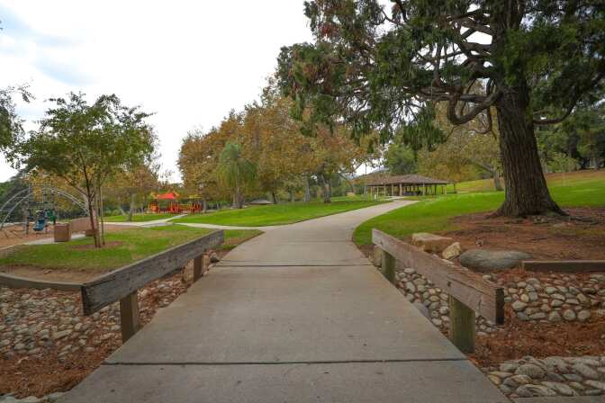 A paved walking trail cuts through grassy picnic areas to the right and a playground to the left. Big trees line the walkway.