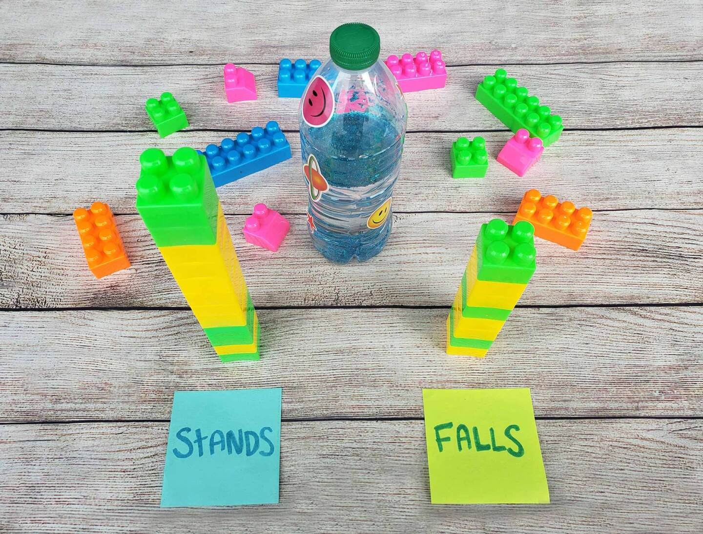 A water bottle with sparkly blue water stands alongside colorful plastic blocks in two towers and two post-its labeled "stands" and "falls."
