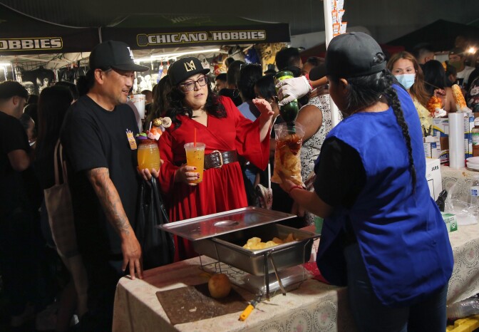 An Asian American man with a baseball cap and a woman in red and a cap order food from a food stall at Avenue 26 night market in Lincoln Heights.