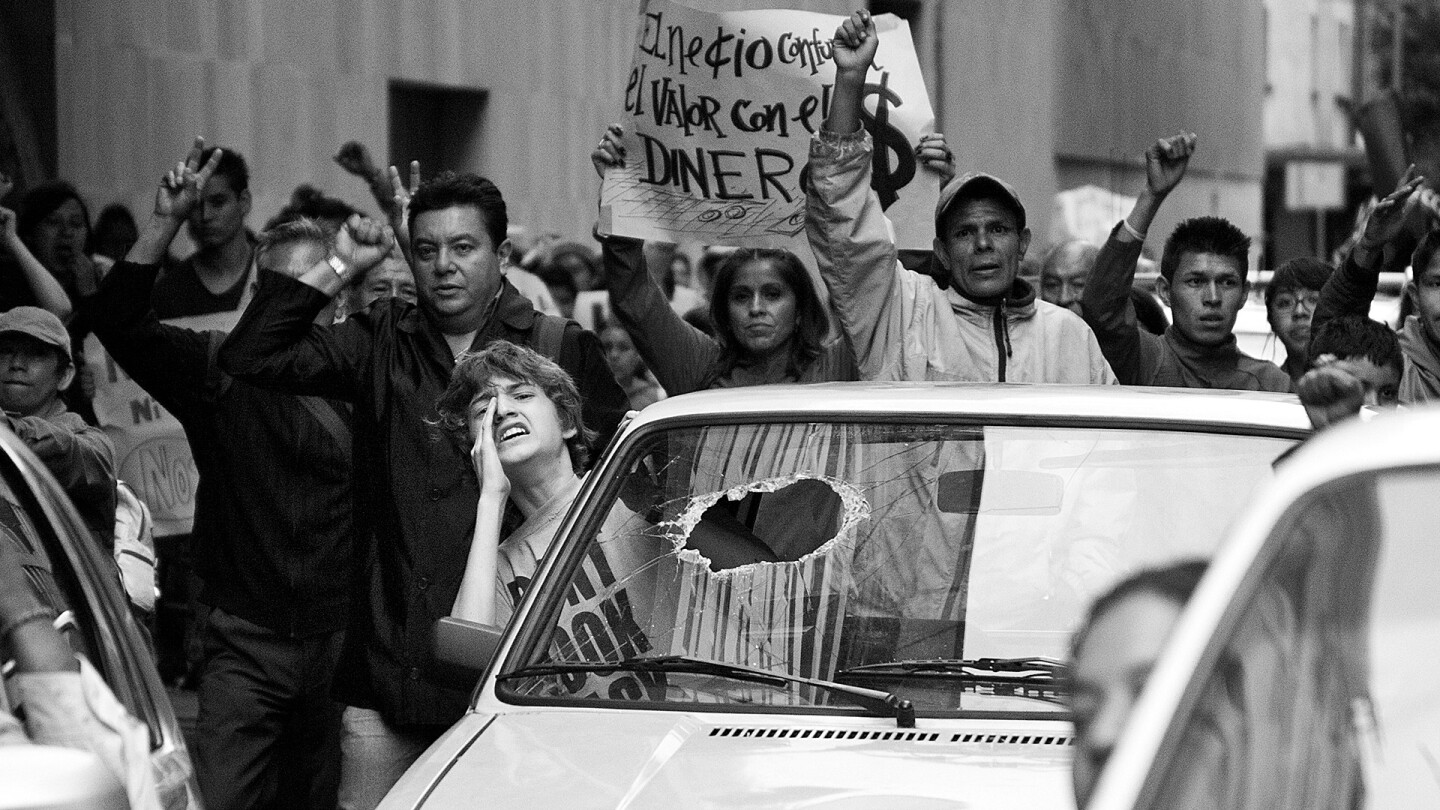 A boy sticks his upper body out a car window and yells out, with a crowd of demonstrators marching behind his vehicle.