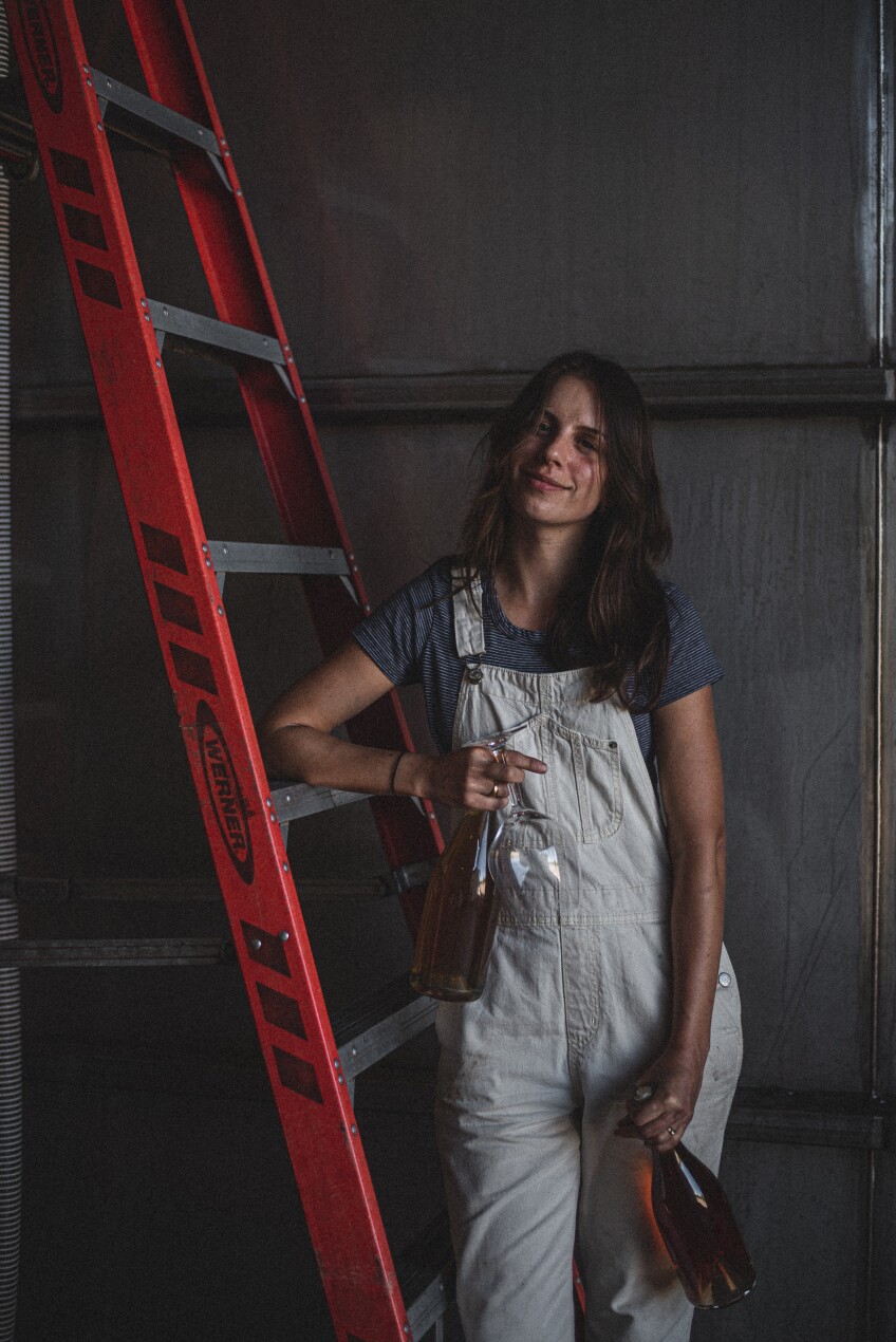 Rachel DeAscentiis leans against a red ladder. She's wearing red overalls and and holds wine glasses in one hand and a bottle of wine in the other.