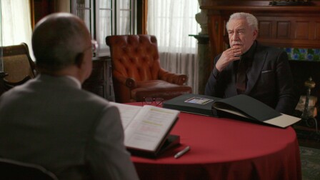 A man with his hand placed on his chin looks at another man sitting across from him at a round table, with a large photobook placed in front of him.
