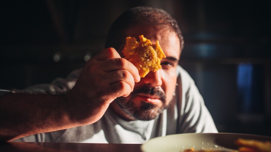 Chef Jose Enrique holds up a fritter over one of his eyes. The plate of fritters can be seen out of focus in the foreground of the photo. He is in a dimly it kitchen with the light source coming from his right.