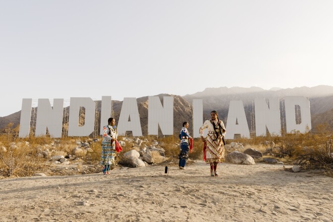 Three women in Indigenous attire dances in front of a large sign in the desert that reads "Indian Land."