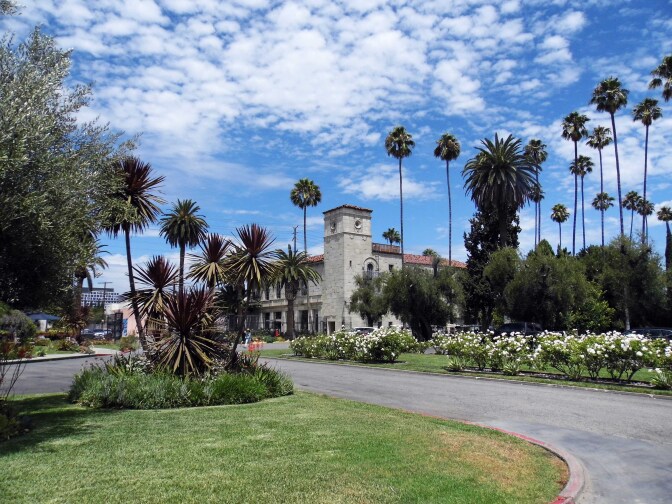 A castle-like building stands before a grassy patch and a paved loop. The building stands among tall palm trees. Above is a bright blue sky with small, fluffy clouds dotted sporadically. 