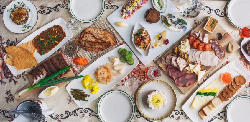 A spread of dishes from Kachka are laid out on a decorative table cloth. The plates and dishes are arranged neatly, perfectly lined up against one another. One of the dishes on the table is a wooden chopping block topped with charcuterie like various cheeses, meats and roasted tomatoes.