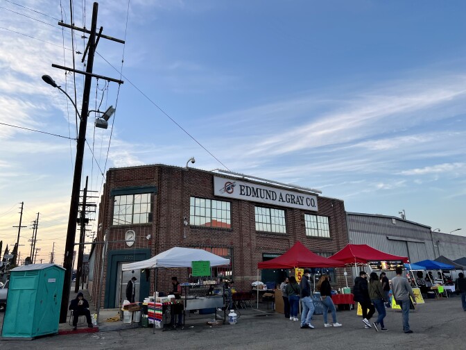 Street vendors under tents line the street in front of a brick building with a sign that reads, "Edmund A. Gray Co." Visitors walk past the tents on the street. Electric lines and a bright blue sky with wispy clouds stretch above the market.