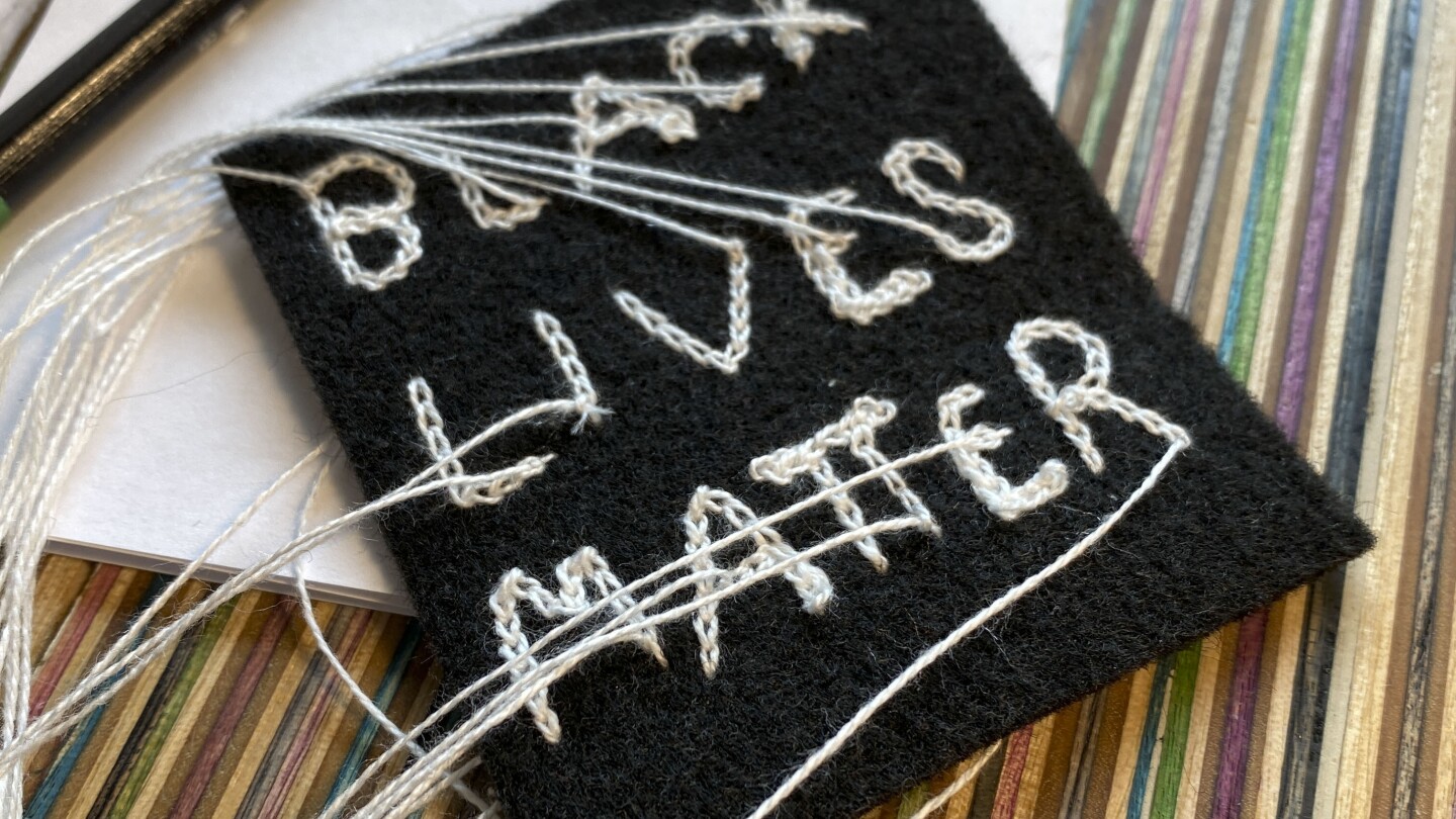 A black, square-shaped piece of fabric with chain stitch embroidery reading the words, "BLACK LIVES MATTER" in white thread. The ends of the threads are long and loose. The patch sits on top of a multicolored, striped surface.