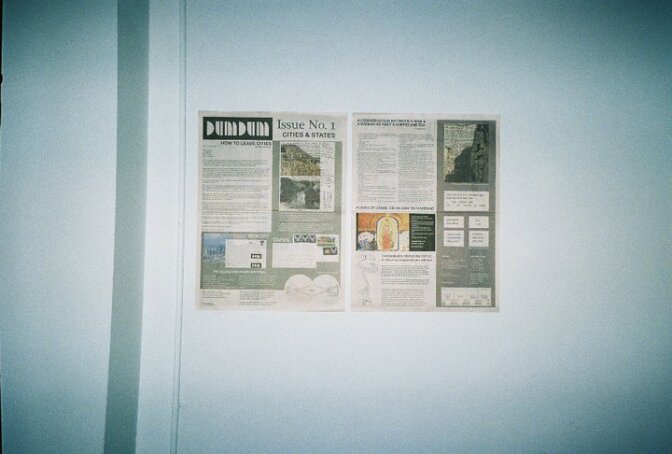 An 11" x 17" newsprint of "Dum Dum Zine" is hanging on a white wall.