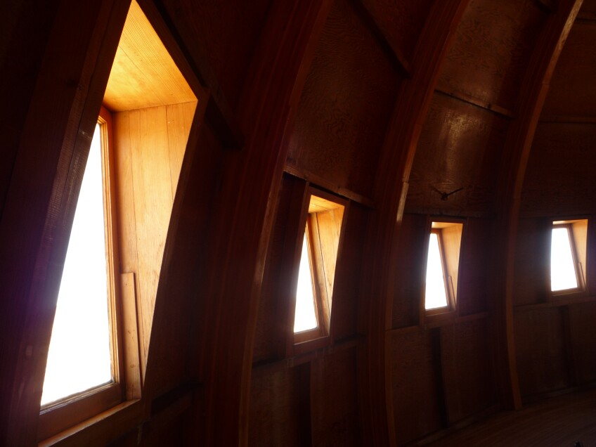 The inside of a dome-shaped building. The inside is dark, with the sunlight coming from rectangular windows partially lighting the room. The inside framing is made of wood. 