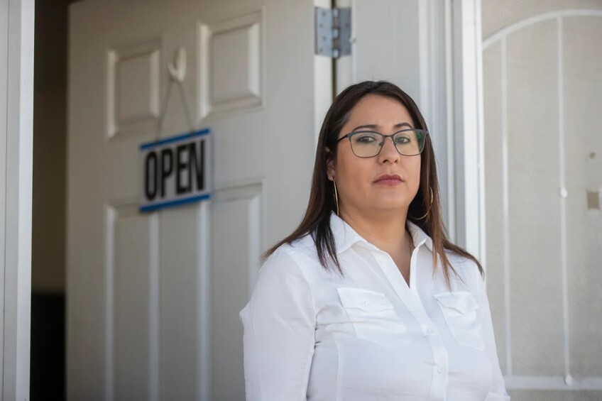 A woman with shoulder-length brown hair wearing glasses, gold hoop earrings and a white buttoned-down shirt stands in front of an open door with on "OPEN" sign hanging on the outside of it.
