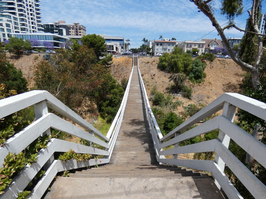 A wooden plank footbridge dips down into a shallow valley and back up onto a residential neighborhood. Apartment complexes and other residential units can be seen from across the valley. On either side of the bridge, brown, dried foliage as well as vibrant green trees and shrubs grow.