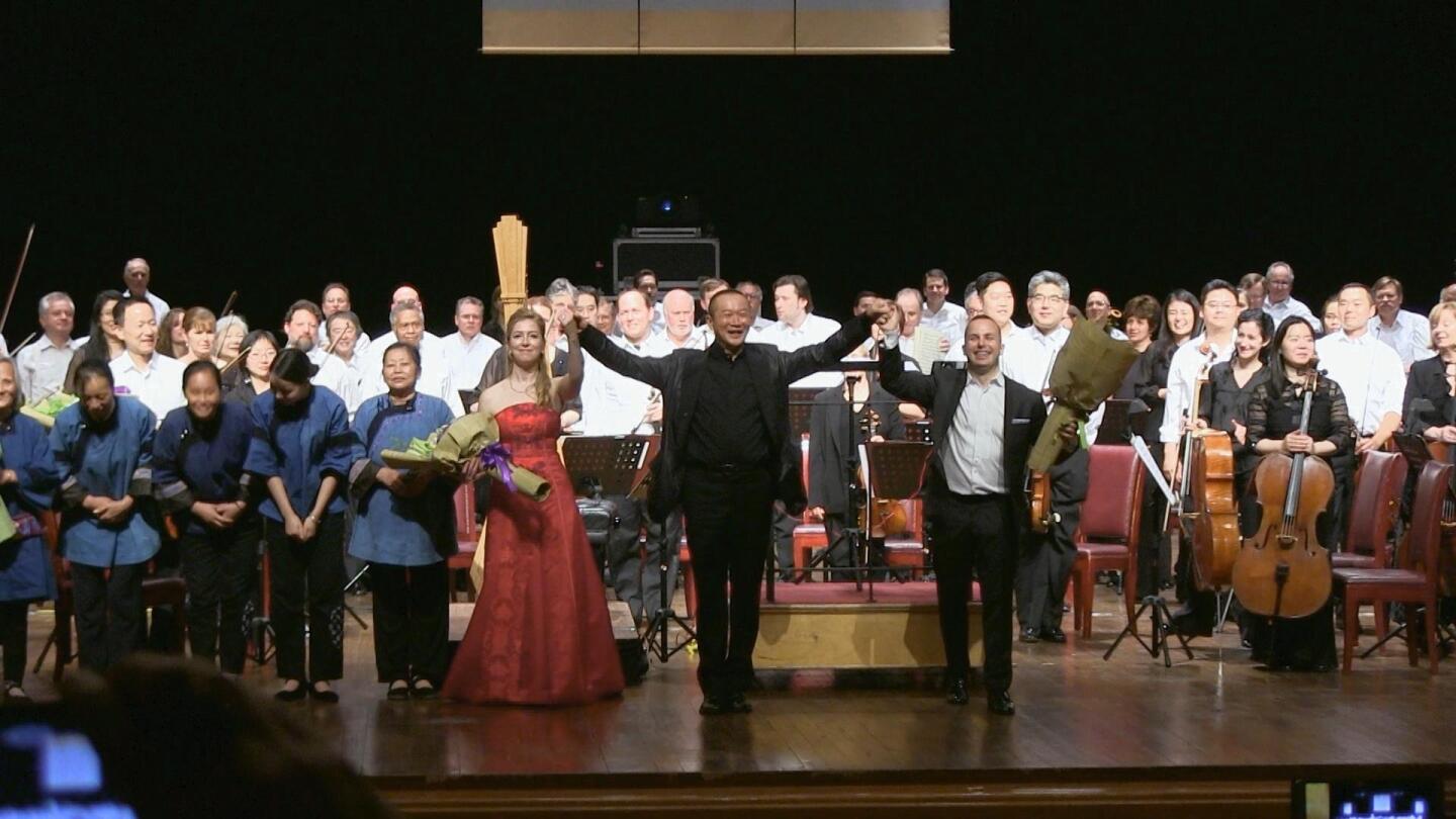 An orchestra and its conductor stand to face the audience in front of them.