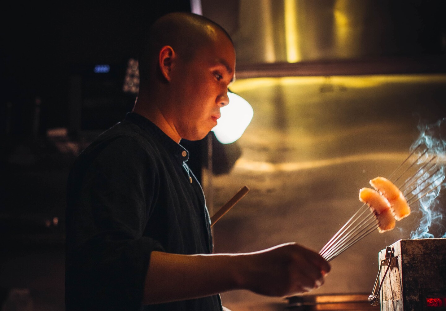 Chef Jon Yao cooks two skewered, bite-sized pieces of fish over a smokey box. He's standing in a dark kitchen and his face is lit by a warm, orange light coming from beneath him.