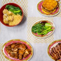 A flat lay photo of Pearl River Deli dishes on a yellow and white checkered tablecloth. In the upper left corner is a bowl of yellow noodles with leafy greens and siu mai. In the lower left corner is a plate of roast poultry in a dark brown sauce. In the middle is a plate of blanched leafy greens with a thick brown sauce drizzled. In the lower right corner is a plate of red char siu pork served over fluffy white rice and a side of leafy vegetables. In the upper right corner is a plate containing a yellow bolo bao with a bone-in breaded pork chop sandwiched between its buns.