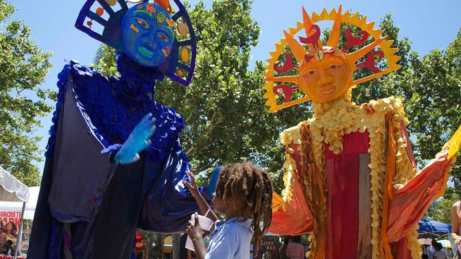 A little boy looks up at two masked figures at the 2017 edition of the Day of the Ancestors: Festival of Masks. | Photo by Sahra Sulaiman, Courtesy of LA Commons