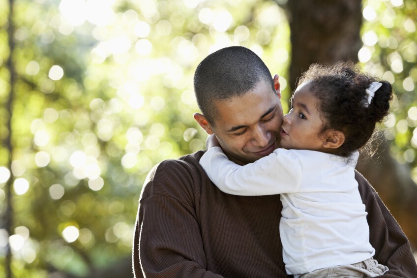 An Hispanic father holding his daughter in his arms on a sunny day at the park. The little girl, 2 years old, has her arms around daddy's neck and is kissing him on the cheek. He is smiling with his eyes closed.