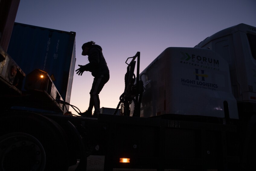 A truck driver stands behind a truck cab in silhouette against a pinkish-blue sky