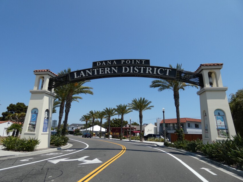 An arch with Spanish-style inspired architecture reads "Dana Point Lantern District" over a black asphalt road. Beyond the arch are palm trees dotted to the side of the street with stores and restaurants lining the road.