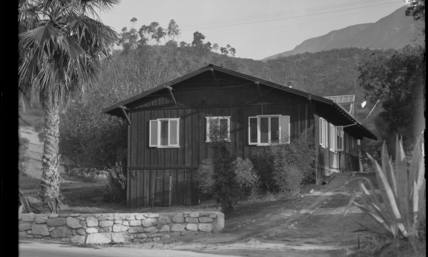 A small wooden, humble building perched on the foothills of a mountain. The building has a plain roof and is comprised of wooden planks all around. Plants can be seen in front of it and a small palm tree to the front.