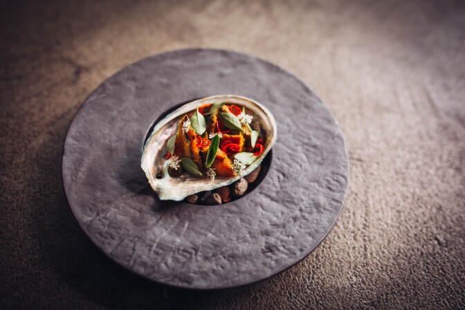 Three cup abalone, sits in a seashell placed atop a round, smooth gray slate. The dish is topped with green leaves and sliced chili.