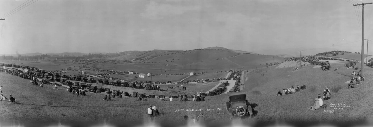 A panoramic view of automobiles parked at the Legion Ascot Speedway in Los Angeles, east of downtown circa 1924. Numerous cars are parked among the low rolling hills, and people are seated and standing amongst the vehicles.