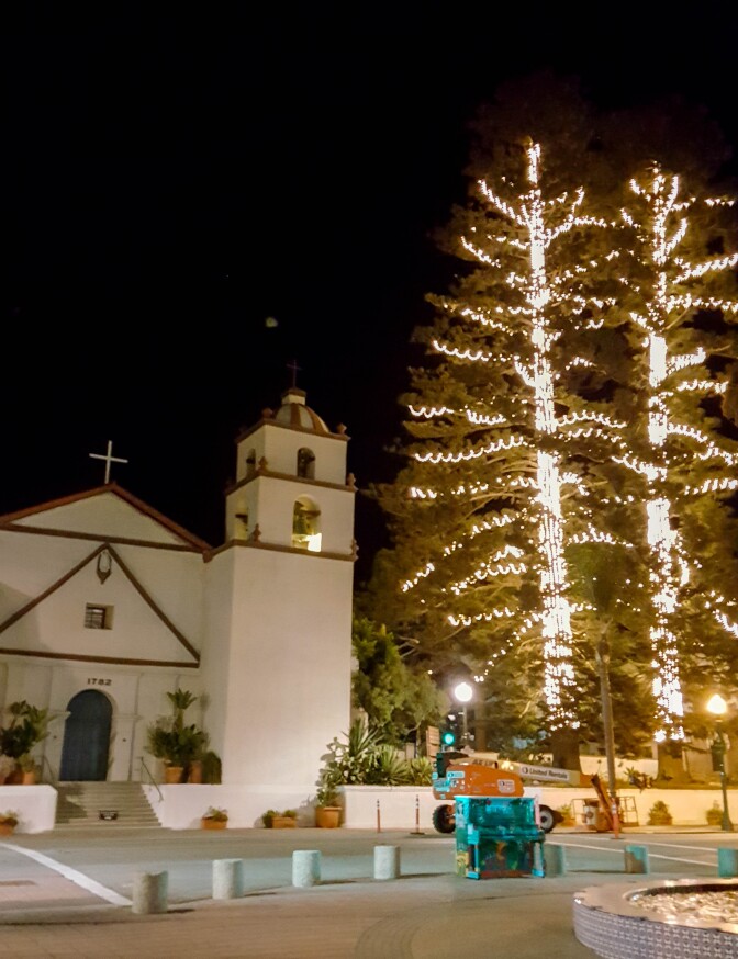 Two tall cedar trees stand by a white church with brown detailing. The trees are lit from the trunk and their various branches.