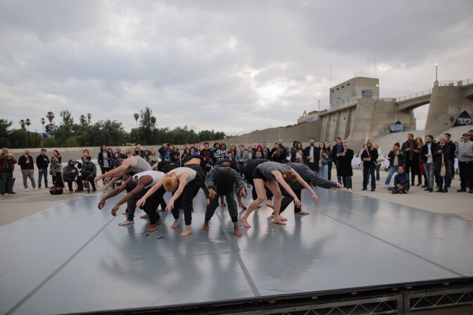 Dancers perform on a gray platform in the middle of a concrete dam. They are arranged in a circle with their backs facing one another. All of the dancers are bent at the waist, with their arms stretched out. Beyond, an audience watches.
