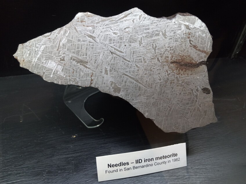 A meteorite with jagged edges and with a chrome, silver finish is on display against a black wooden surface and is propped up on a clear acrylic stand.