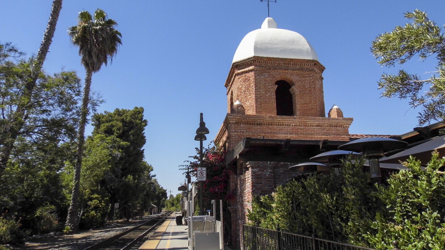 A Mission Revival style red brick building stands next to a set of train tracks that cut between the station platform and a line of trees.