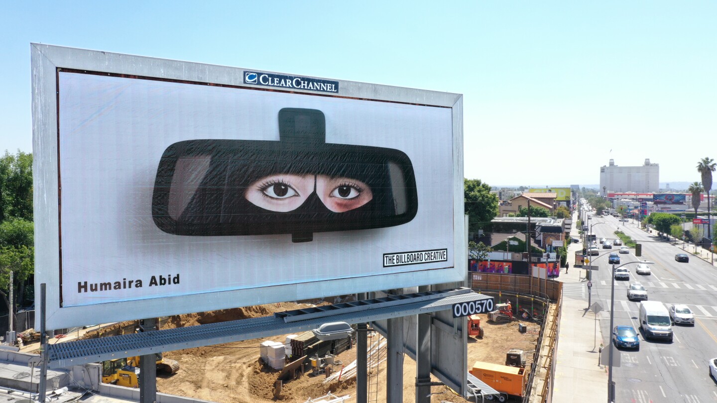 A billboard in L.A. shows a woman's reflection in a car's rear view mirror. Wearing a niqab, the woman's eyes are the only visible part of her face, and one eye bears a noticeable bruise. Below is a busy street with cars bustling by.