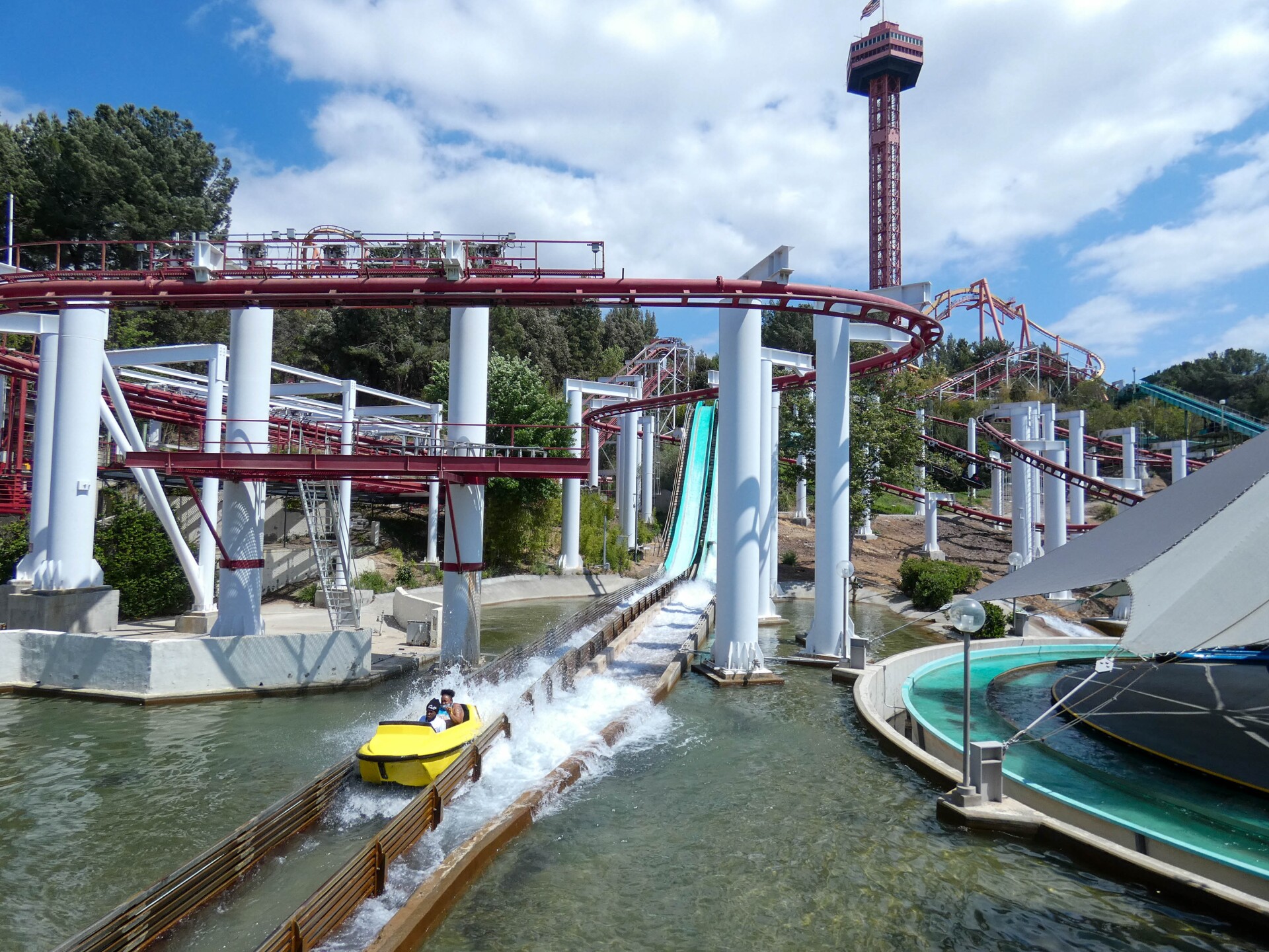 Two passengers on a yellow ride car glide down a river stream ramp at the end of a flume ride. Water sprays up behind them as the splash ride car zooms through a pool of water. Behind the riders are elaborate coaster tracks and river streams from various rides looped and tangled with one another. A tall red view tower towers behind everything in the near distance.