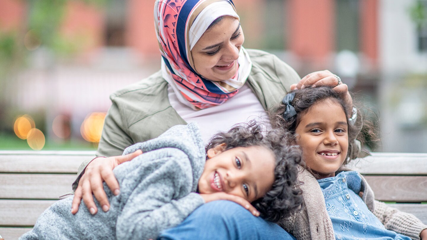 A smiling woman wearing a hijab sits with two smiling children on a bench. | stock