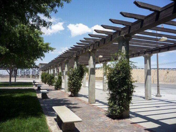 Pergolas line a walkway, offering partial shade to the path. Foliage creep up the columns holding up the pergola. To the left of the walkway is dark green, healthy grass partially shaded by tree branches that are peeking into the top left corner of the photo.